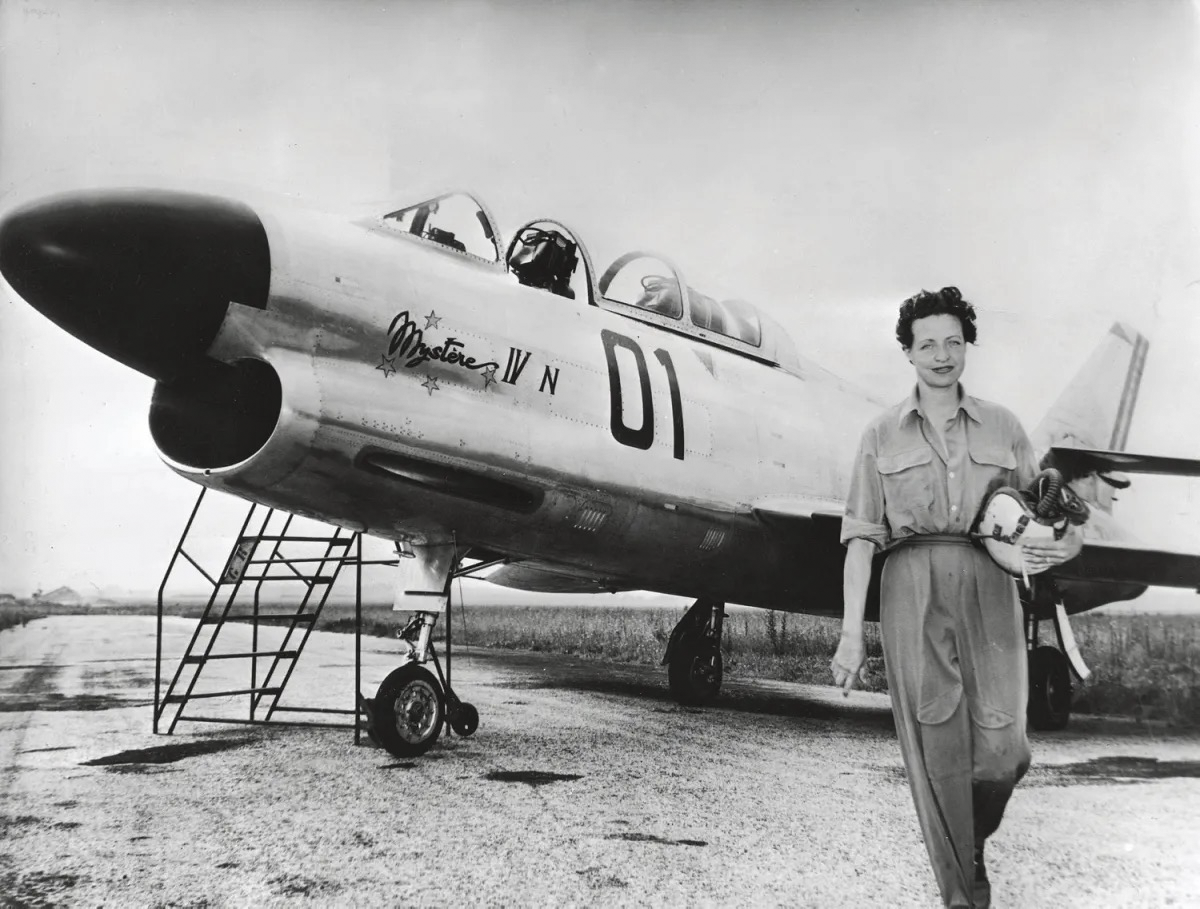 acqueline Auriol, aviation pioneer, in a flight suit holding her helmet, walking in front of the Dassault Mystère IV N jet on a tarmac.